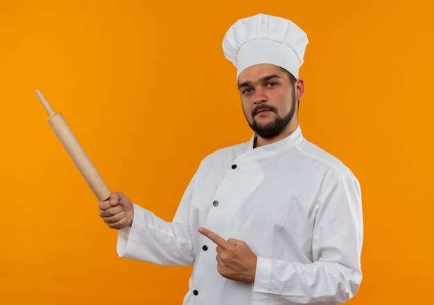 Confiant jeune homme cuisinier en uniforme de chef tenant un rouleau à pâtisserie et pointant sur le côté isolé sur un mur orange