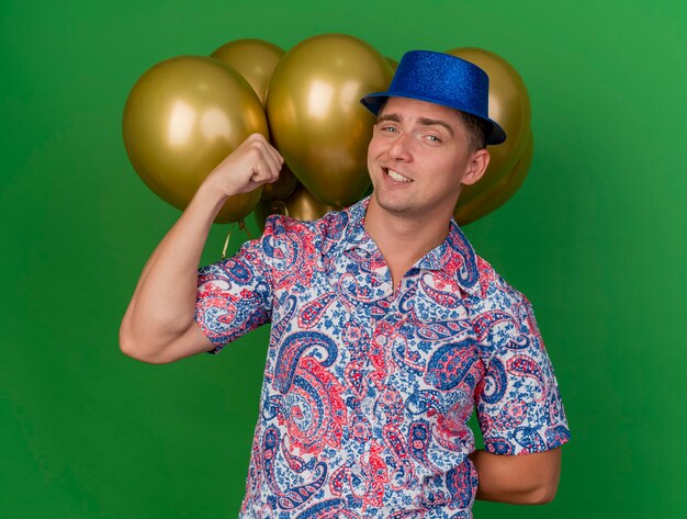Confiant jeune homme de fête portant un chapeau bleu debout devant des ballons et montrant oui geste isolé sur vert
