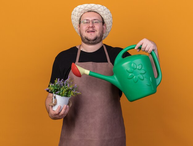 Confiant jeune homme jardinier portant un chapeau de jardinage et des gants tenant un arrosoir avec une fleur dans un pot de fleurs isolé sur un mur orange