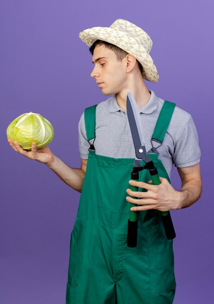 Confiant jeune homme jardinier portant un chapeau de jardinage tient des tondeuses et regarde le chou