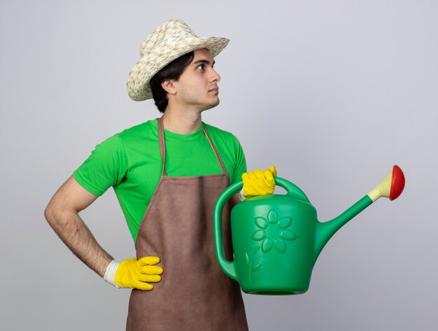 Confiant jeune homme jardinier en uniforme portant chapeau de jardinage à côté tenant arrosage peut mettre la main sur la hanche
