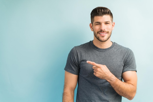 Confiant jeune homme latin portant un t-shirt gris et pointant vers un espace vide tout en établissant un contact visuel sur fond uni