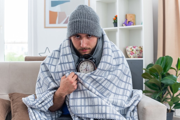 Confiant Jeune Homme Malade Portant Une écharpe Et Un Chapeau D'hiver Assis Sur Un Canapé Dans Le Salon Enveloppé Dans Une Couverture Tenant Un Réveil Regardant à L'avant