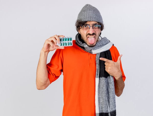 Confiant jeune homme malade portant des lunettes chapeau d'hiver et écharpe tenant et pointant sur pack de capsules médicales montrant la langue à l'avant isolé sur mur blanc