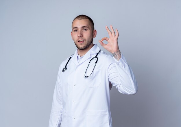 Confiant jeune homme médecin portant une robe médicale et un stéthoscope autour de son cou faisant signe ok isolé sur blanc avec espace copie