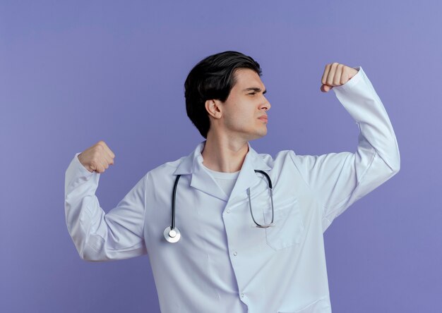 Confiant jeune homme médecin portant une robe médicale et un stéthoscope faisant un geste fort regardant le poing isolé sur mur violet