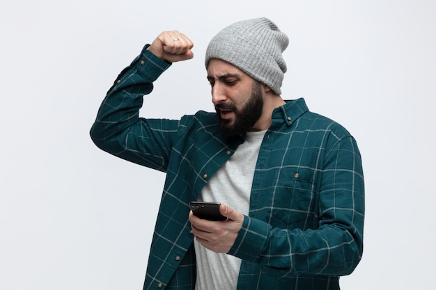Confiant jeune homme portant un chapeau d'hiver tenant et regardant un téléphone portable montrant un geste fort isolé sur fond blanc