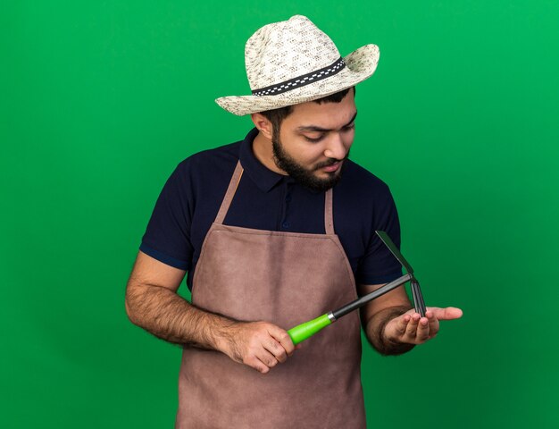 Confiant jeune homme de race blanche jardinier portant chapeau de jardinage tenant et regardant houe râteau isolé sur mur vert avec espace copie