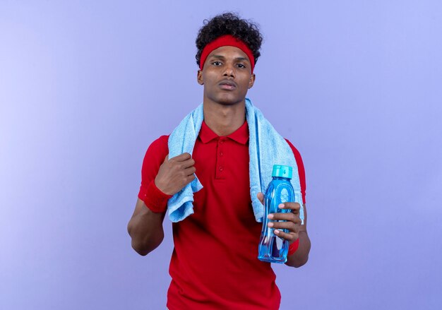 Confiant jeune homme sportif afro-américain portant un bandeau et un bracelet tenant une bouteille d'eau avec une serviette sur l'épaule