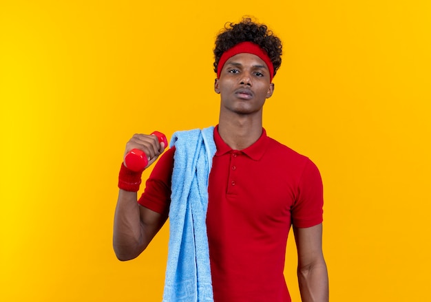 Confiant jeune homme sportif afro-américain portant bandeau et bracelet tenant un haltère avec une serviette sur l'épaule isolé sur mur jaune