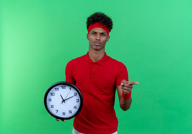 Confiant jeune homme sportif afro-américain portant un bandeau et un bracelet tenant une horloge murale et vous montrant le geste isolé sur fond vert