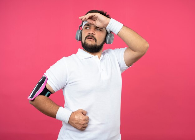 Confiant jeune homme sportif portant un bandeau et un bracelet avec des écouteurs et un brassard de téléphone mettant la main sur le front isolé sur un mur rose