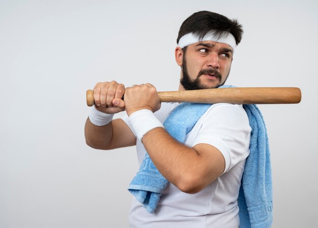 Confiant jeune homme sportif regardant côté portant un bandeau et un bracelet avec une serviette sur l'épaule tenant une batte de baseball sur l'épaule isolé sur un mur blanc avec espace de copie