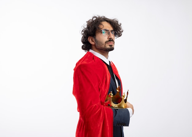 Confiant jeune homme de super-héros caucasien dans des lunettes optiques portant un costume avec une cape rouge se tient sur le côté tenant une couronne