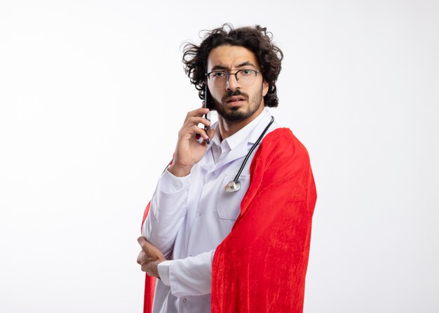 Confiant jeune homme de super-héros caucasien à lunettes optiques portant l'uniforme du médecin avec manteau rouge et avec stéthoscope autour du cou se tient sur le côté en parlant au téléphone