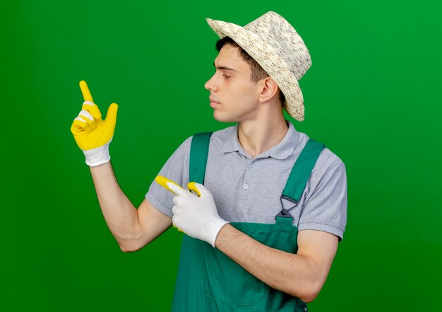 Confiant jeune jardinier mâle portant chapeau et gants de jardinage regarde et pointe sur le côté