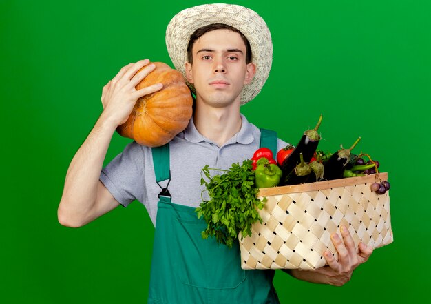 Photo gratuite confiant jeune jardinier mâle portant chapeau de jardinage détient panier de légumes et à la citrouille