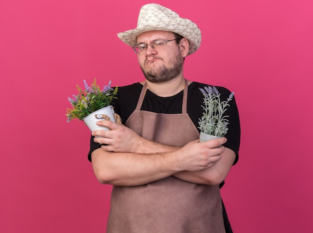 Confiant jeune jardinier mâle portant chapeau de jardinage tenant et traversant des fleurs dans des pots de fleurs isolés sur un mur rose