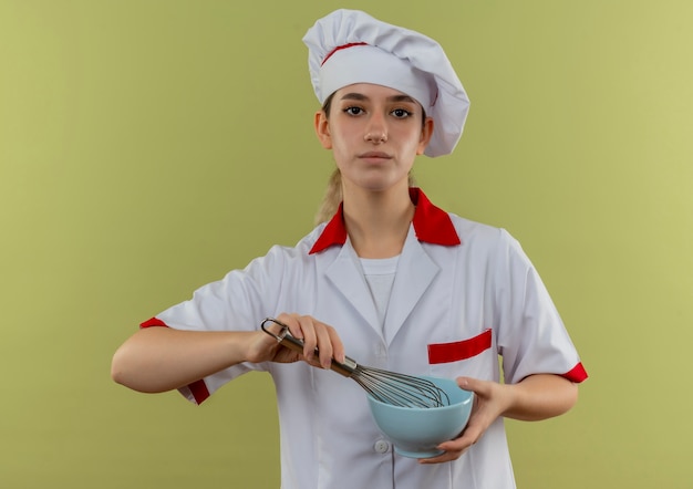 Confiant Jeune Joli Cuisinier En Uniforme De Chef Tenant Un Fouet Et Un Bol Isolé Sur Un Mur Vert