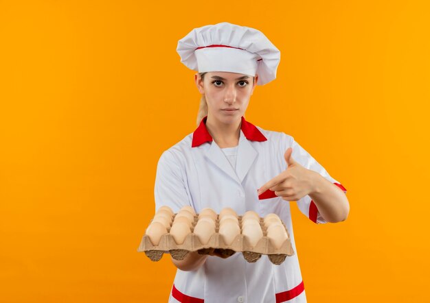 Confiant jeune joli cuisinier en uniforme de chef tenant et pointant sur un carton d'œufs isolé sur un mur orange avec espace de copie