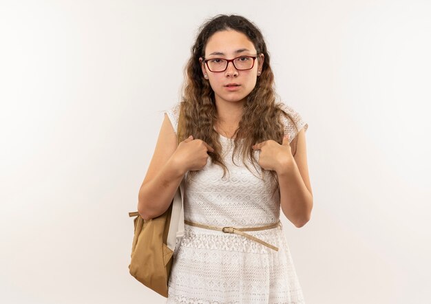 Confiant jeune jolie écolière portant des lunettes et sac à dos pointant sur elle-même isolé sur blanc avec copie espace