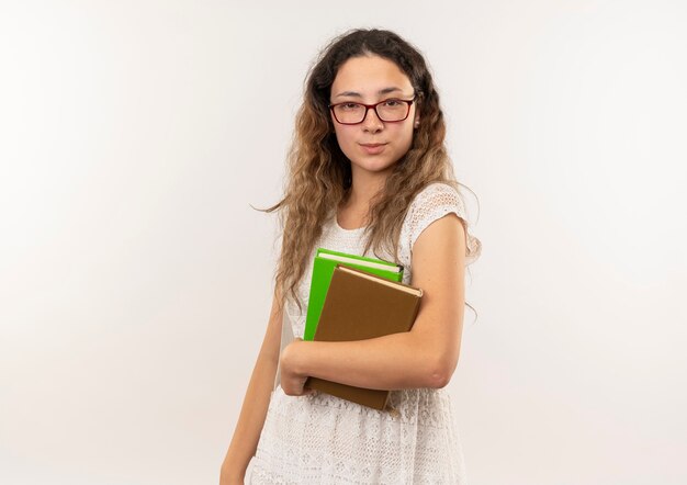 Confiant jeune jolie écolière portant des lunettes et sac à dos tenant des livres à isolé sur blanc avec espace copie