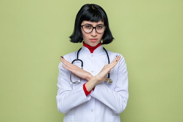 Confiant jeune jolie femme de race blanche avec des lunettes en uniforme de médecin avec stéthoscope croisant les mains ne faisant aucun signe