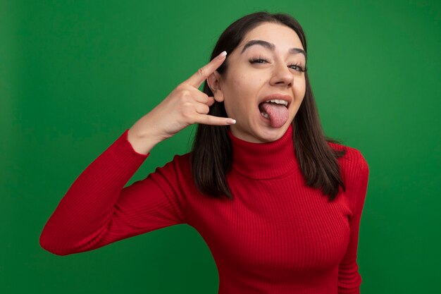 Confiant jeune jolie fille caucasienne gardant la main près de la tête faisant rock signe montrant la langue isolé sur mur vert