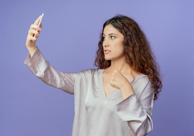 Confiant jeune jolie fille prendre un selfie et tenant le poing isolé sur mur bleu