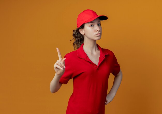 Confiant jeune jolie livreuse en uniforme rouge et capuchon mettant la main sur la taille et levant le doigt isolé sur fond orange avec espace de copie