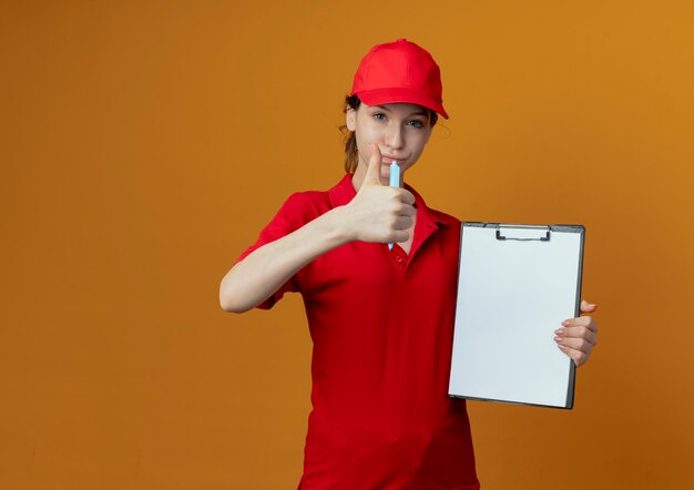 Confiant jeune jolie livreuse en uniforme rouge et capuchon tenant stylo et presse-papiers et montrant le pouce vers le haut à la caméra isolée sur fond orange avec espace de copie