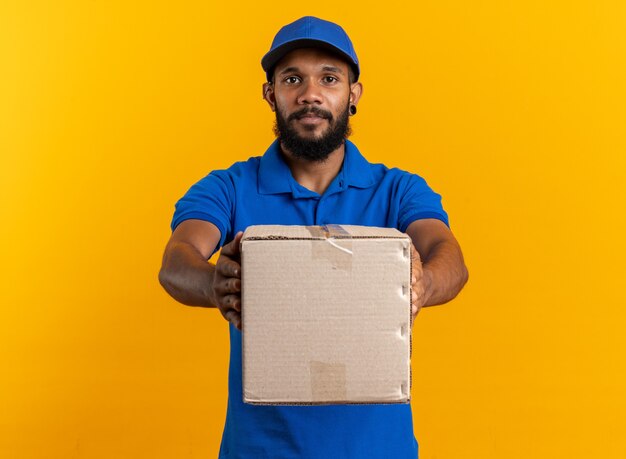 Confiant jeune livreur afro-américain tenant une boîte en carton isolée sur fond orange avec espace de copie