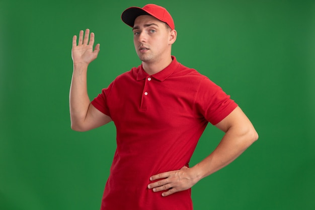 Confiant jeune livreur en uniforme et casquette montrant bonjour geste mettant la main sur la hanche isolé sur mur vert