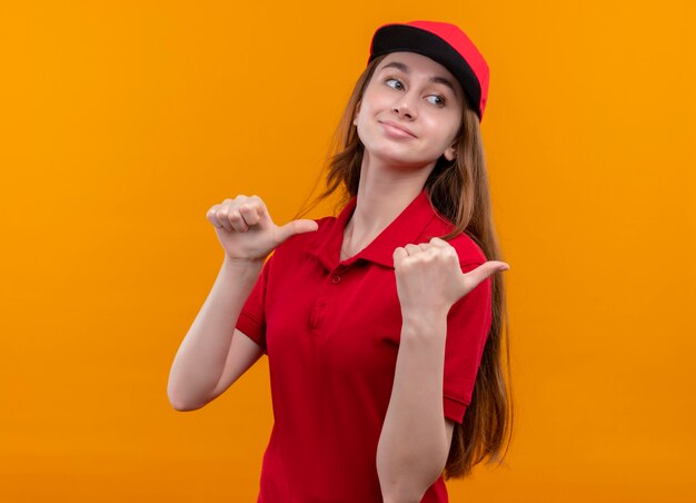 Confiant jeune livreuse en uniforme rouge montrant les pouces vers le haut à droite sur l'espace orange isolé avec copie espace