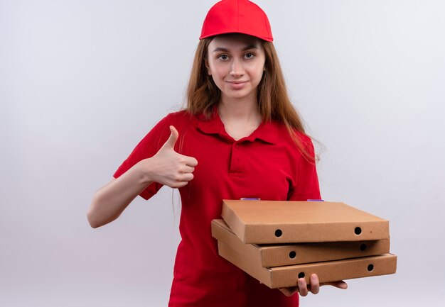 Confiant jeune livreuse en uniforme rouge tenant des paquets et montrant le pouce vers le haut sur un espace blanc isolé