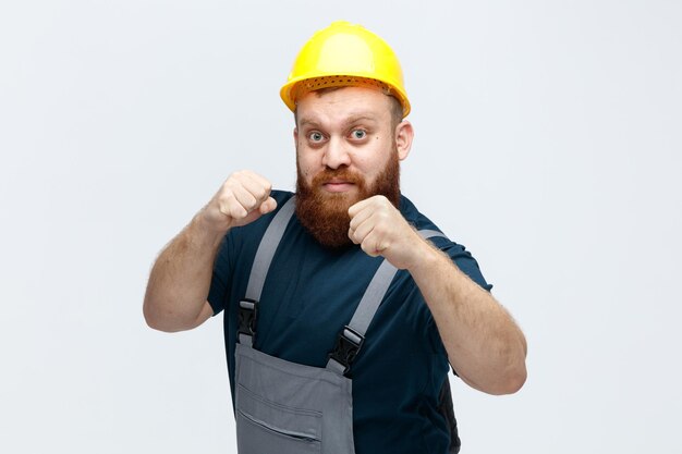 Confiant jeune ouvrier du bâtiment masculin portant un casque de sécurité et un uniforme regardant la caméra montrant un geste de boxe isolé sur fond blanc