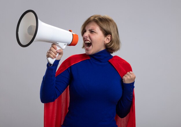 Confiant jeune superwoman blonde en cape rouge criant dans le haut-parleur serrant le poing à côté isolé sur mur blanc