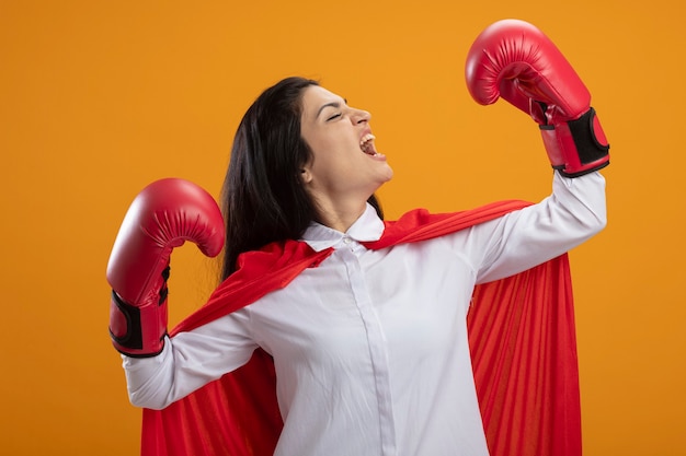 Confiant jeune superwoman portant des gants de boîte faisant un geste fort tournant la tête à l'autre en criant avec les yeux fermés isolé sur le mur orange