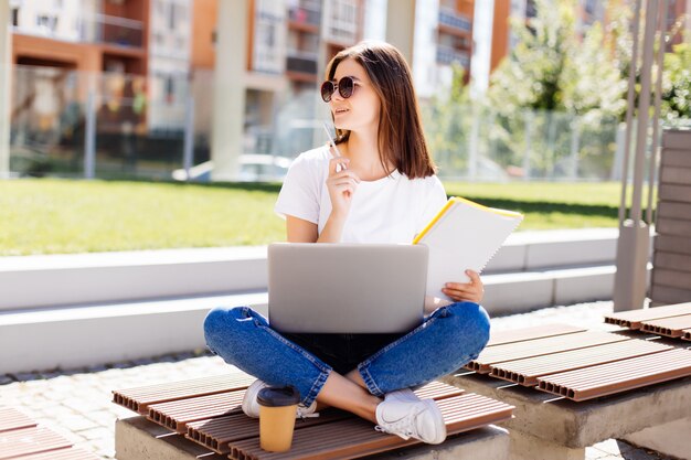 Confiant jolie femme assise sur un banc dans le parc et la messagerie dans les médias sociaux lors de l'écriture dans le cahier pendant la pause-café