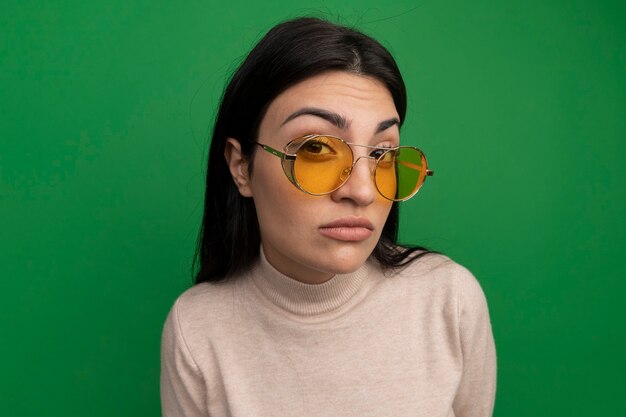 Confiant jolie femme brune à lunettes de soleil regarde à l'avant isolé sur mur vert