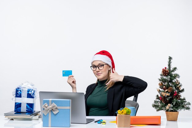 Confiant jolie femme avec chapeau de père Noël et portant des lunettes assis à une table et tenant une carte bancaire et faisant appelez-moi geste au bureau