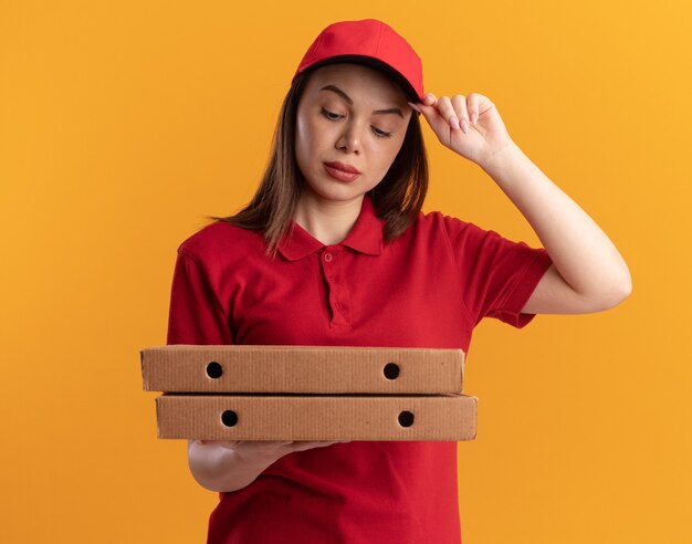 Confiant jolie femme de livraison en uniforme met la main sur le chapeau tenant et regardant des boîtes de pizza sur orange