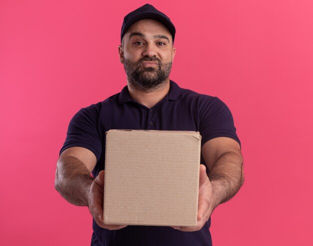 Confiant livreur d'âge moyen en uniforme et cap tenant la boîte à l'avant isolé sur mur rose