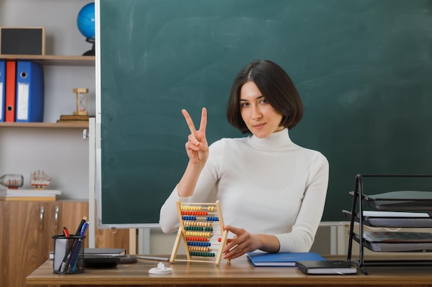 confiant montrant un geste de paix jeune enseignante tenant un boulier assis au bureau avec des outils scolaires en classe