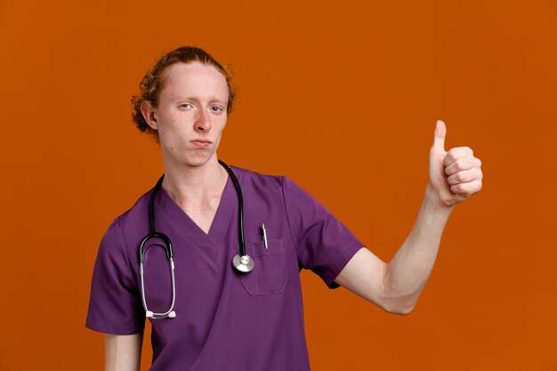 confiant montrant les pouces vers le haut jeune homme médecin portant l'uniforme avec stéthoscope isolé sur fond orange