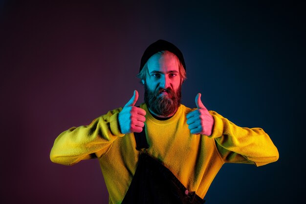 Confiant montrant les pouces vers le haut. Portrait de l'homme caucasien sur fond de studio dégradé en néon. Beau modèle masculin avec un style hipster. Concept d'émotions humaines, expression faciale, ventes, publicité.