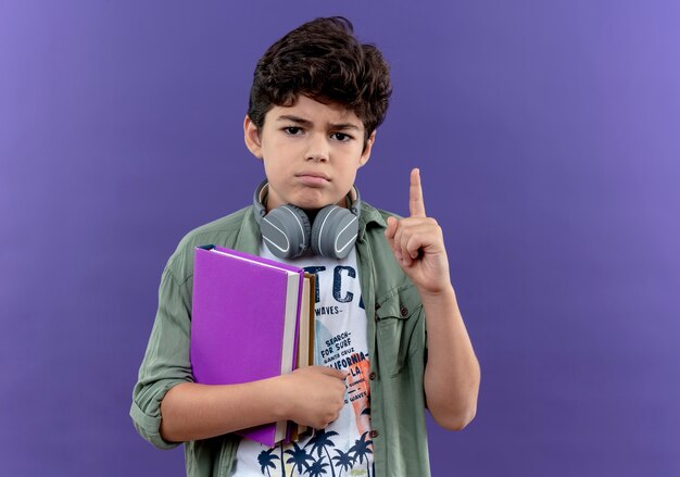 Confiant petit écolier portant des écouteurs tenant des livres et des points en haut