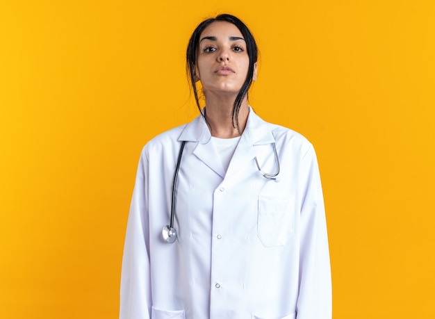 Confiant en regardant la caméra jeune femme médecin portant une robe médicale avec stéthoscope isolé sur mur jaune