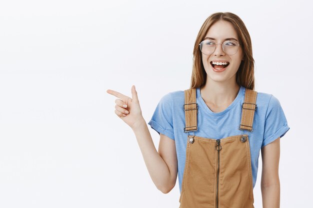 Confiant souriant étudiant jolie fille dans des verres à la recherche de plaisir, pointant le doigt à gauche sur votre logo