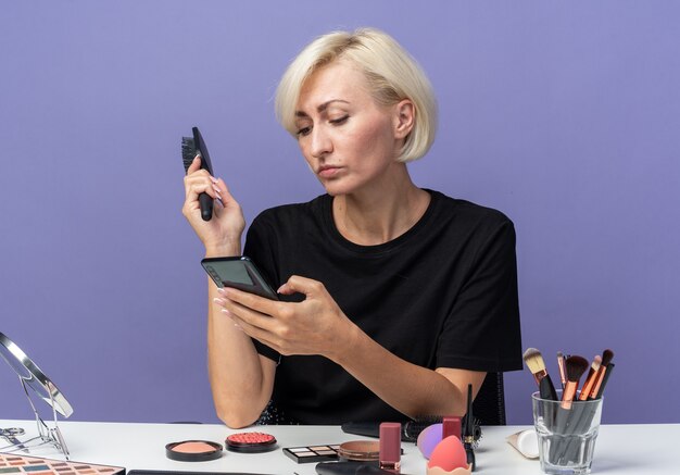 Confiante, belle jeune fille assise à table avec des outils de maquillage tenant un peigne et regardant le téléphone dans sa main isolée sur un mur bleu